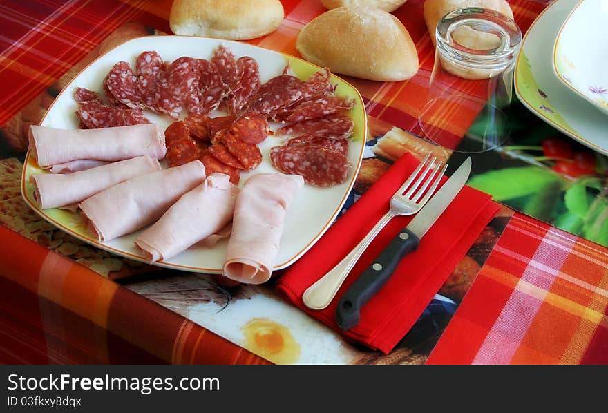 Piece of a laid table with a plate full of sliced salami and ham, cutlery, a glass and some bread.