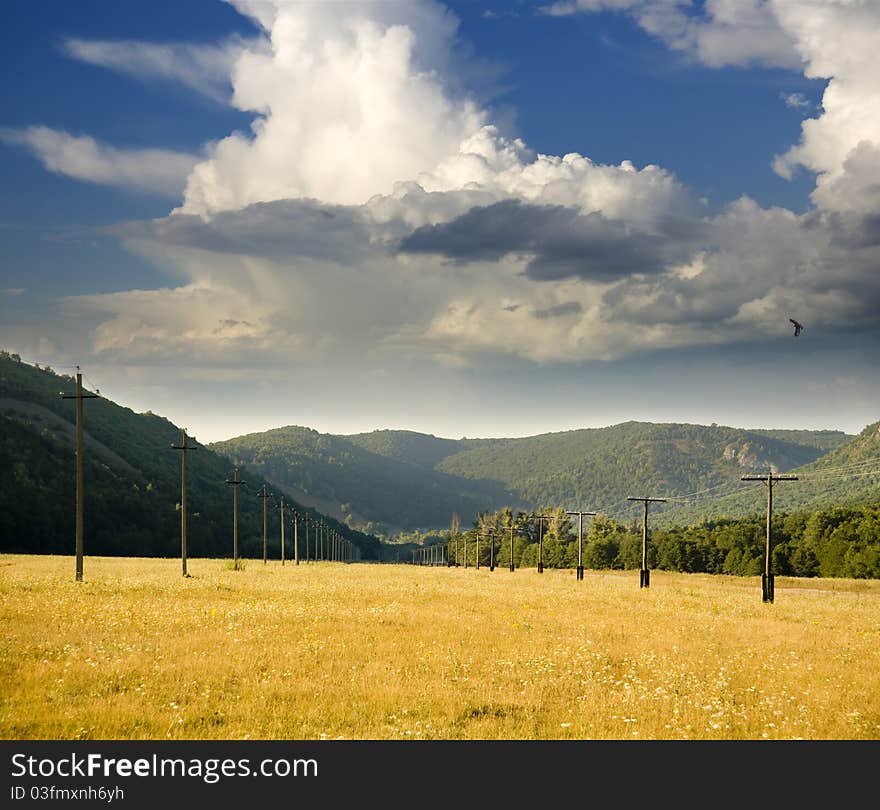 Power line is located in a mountain valley in the pasture with dry grass on a background of white large clouds. Power line is located in a mountain valley in the pasture with dry grass on a background of white large clouds.