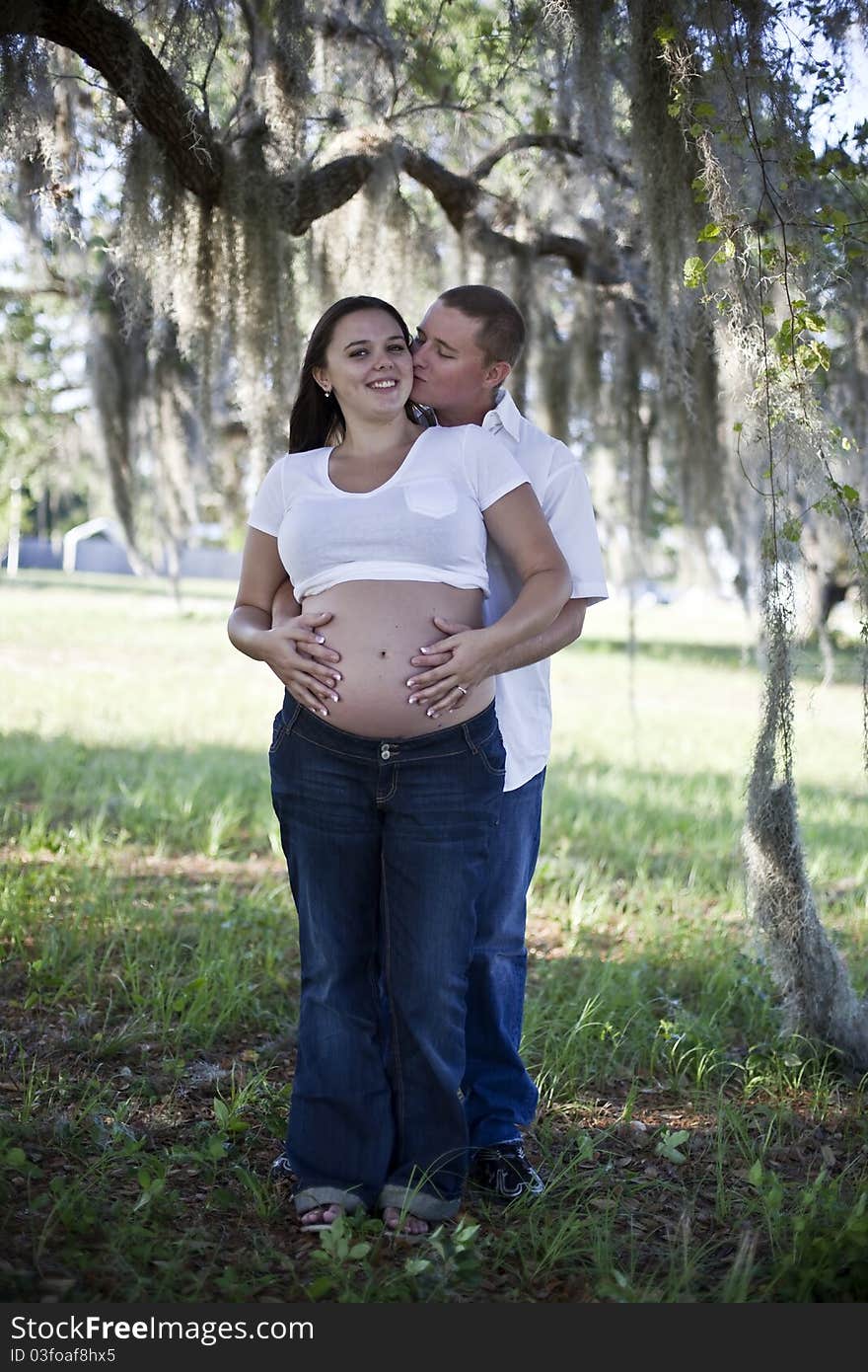 A young man kissing the cheek of a pregnant woman outside under a tree. A young man kissing the cheek of a pregnant woman outside under a tree.