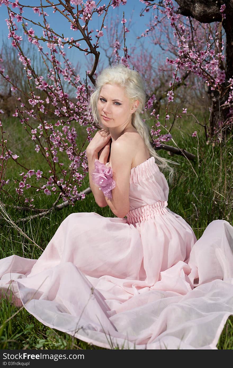 Outdoor shot of Young pretty blond woman wearing pink dress sitting on the grass in blooming garden. Outdoor shot of Young pretty blond woman wearing pink dress sitting on the grass in blooming garden