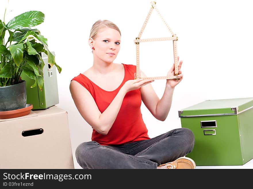 Young woman with a folding rule as a house symbol sits between move cardboards and a plant. Young woman with a folding rule as a house symbol sits between move cardboards and a plant