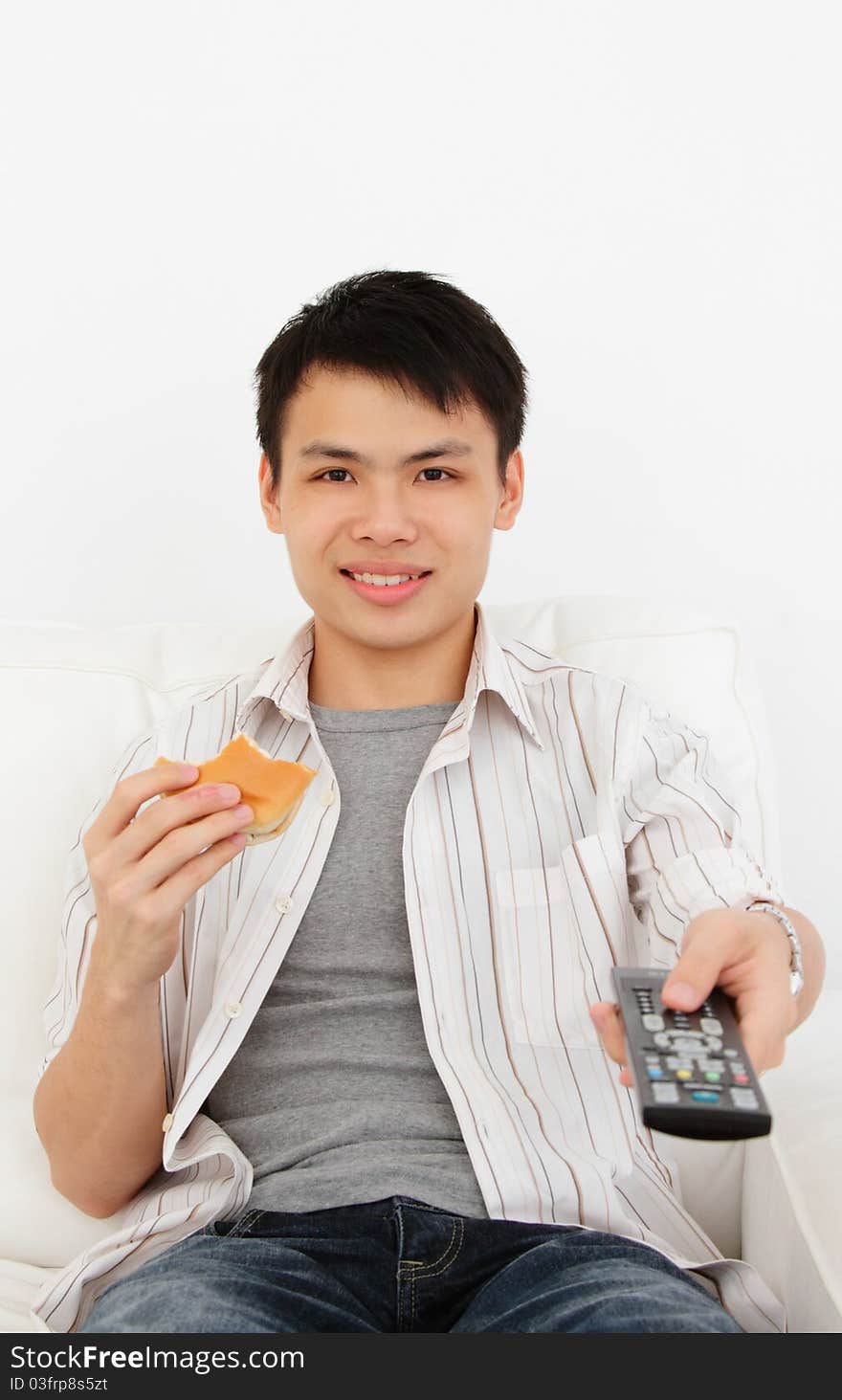 A young Asian man with a TV remote and burger on a sofa against a white background. A young Asian man with a TV remote and burger on a sofa against a white background