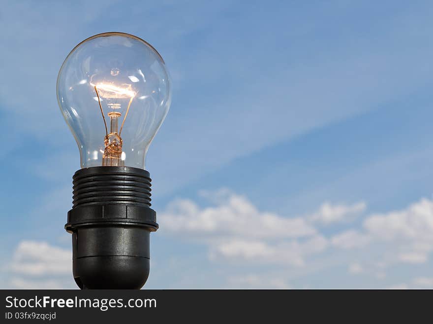 Clear electric light bulb glowing against blue sky and puffy cloud background