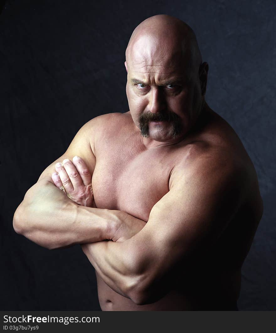 A bald headed body builder with arms crossed glaring at the camera