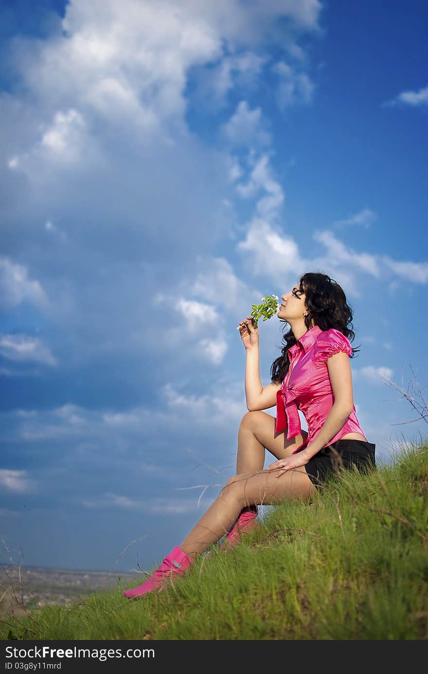 Beautiful girl in pink blouse and short skirt sitting at the top of the hill on a background of blue sky and sniffs a flower. Beautiful girl in pink blouse and short skirt sitting at the top of the hill on a background of blue sky and sniffs a flower.