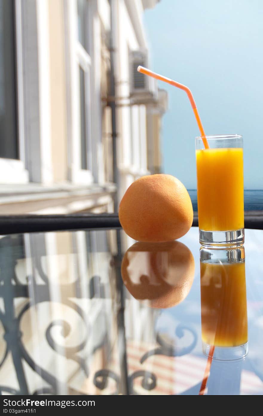 Glass of orange juice with a tubule. A glass table. The sea on horizon.