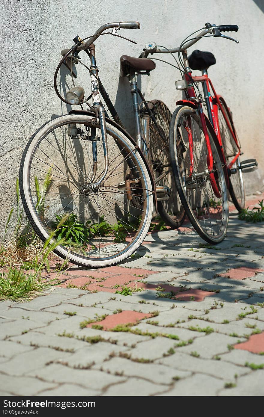 Two bikes, one blue, the other red are leaning against the wall on a sunny day. Two bikes, one blue, the other red are leaning against the wall on a sunny day