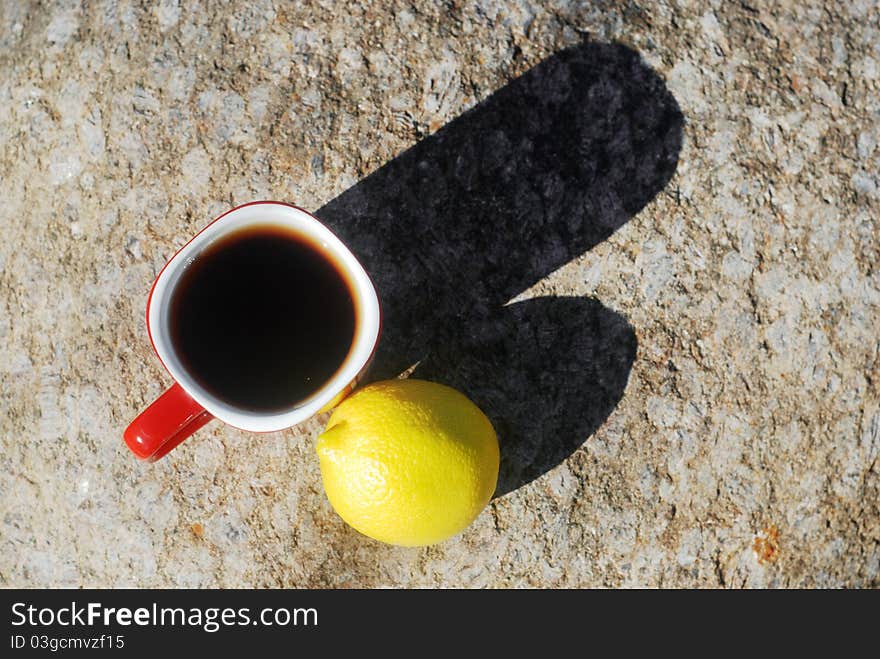 Red cup of black tea and a whole lemon fruit close-up on sunny day with long shadows outdoors. Stone background, view from above. Red cup of black tea and a whole lemon fruit close-up on sunny day with long shadows outdoors. Stone background, view from above