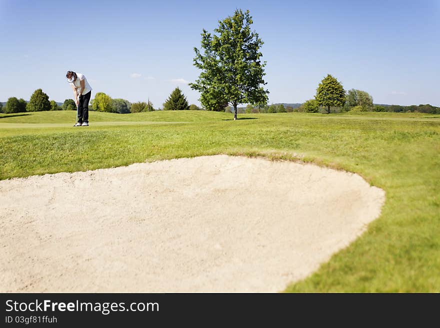 Active senior female golf player putting golf ball on green on beautiful golf course with sand bunker in foreground. Active senior female golf player putting golf ball on green on beautiful golf course with sand bunker in foreground.