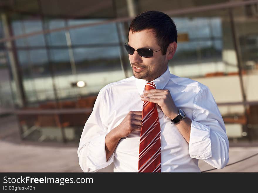 Close up of confident businessman in white shirt adjusting his orange tie, standing outdoors in front of glass office building. Close up of confident businessman in white shirt adjusting his orange tie, standing outdoors in front of glass office building.