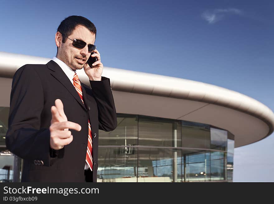 Young businessman in black suit and sunglasses being busy on cell phone with office building and blue sky in background. Young businessman in black suit and sunglasses being busy on cell phone with office building and blue sky in background.