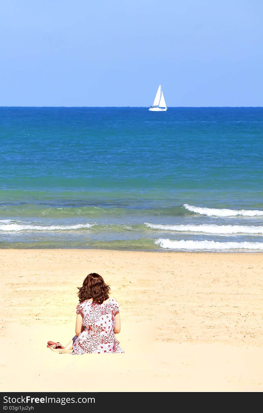 Girl sitting on a sandy beach and looking at a small white sail in the blue sea. Girl sitting on a sandy beach and looking at a small white sail in the blue sea