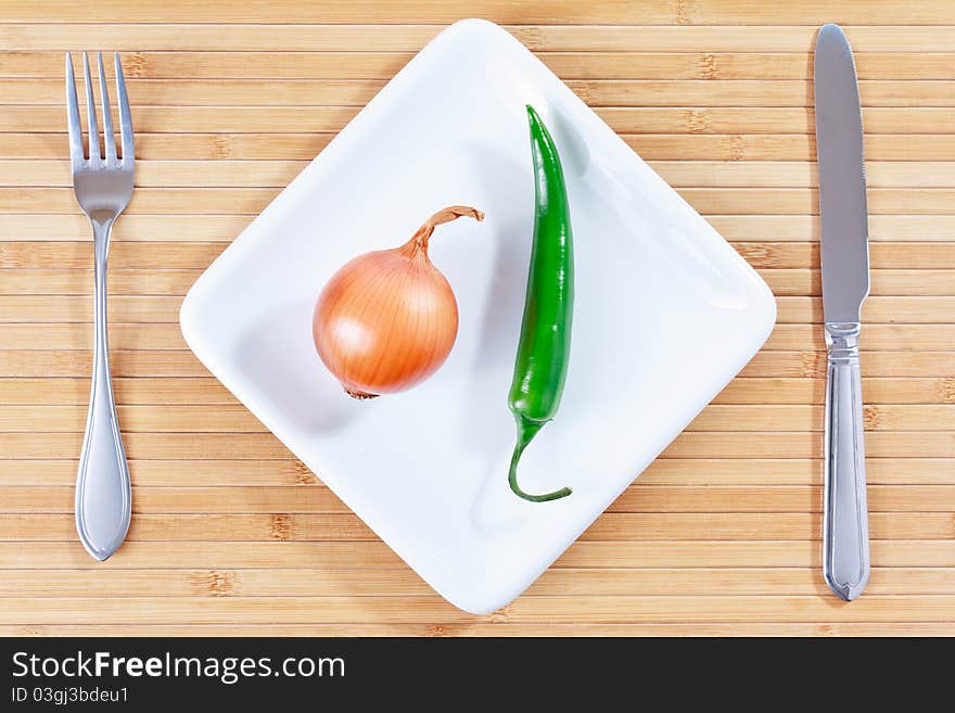 Green chili pepper and onion on a white plate and bamboo placemat with fork and knife