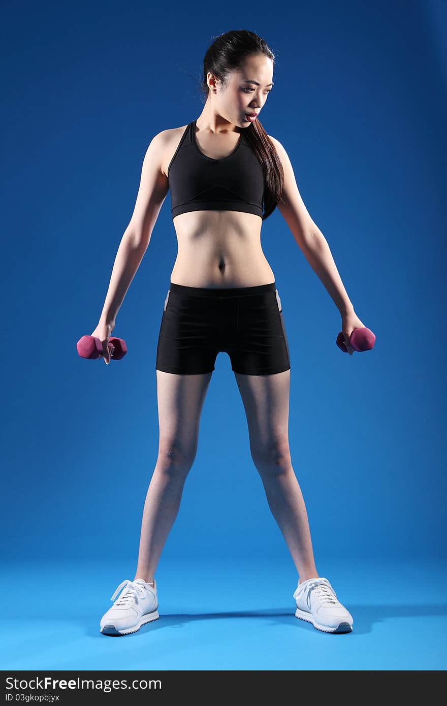 Beautiful young Asian Chinese girl working out with hand weights, wearing black sports bra and shorts, standing in blue studio. Beautiful young Asian Chinese girl working out with hand weights, wearing black sports bra and shorts, standing in blue studio.