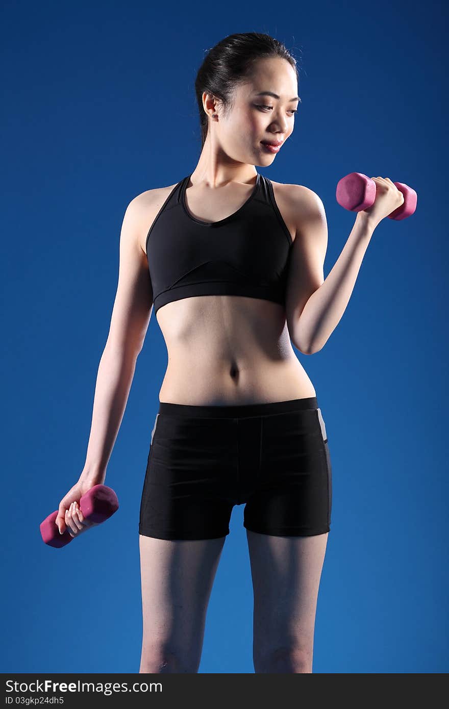 Beautiful young oriental woman working out with fitness hand weights, wearing black sports bra and shorts, standing in blue studio. Beautiful young oriental woman working out with fitness hand weights, wearing black sports bra and shorts, standing in blue studio.