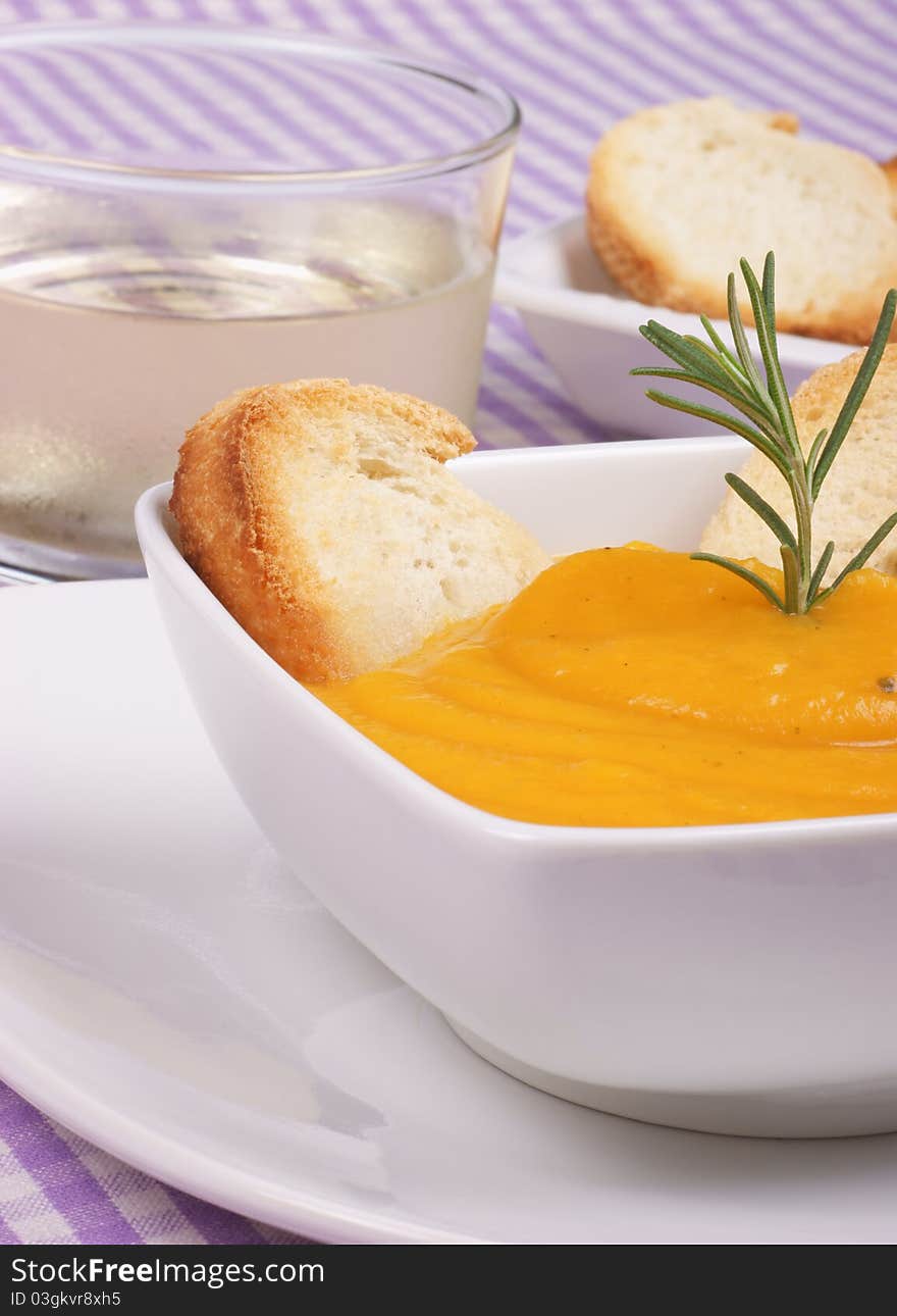 Cream of squash soup in a white plate with toasted bread and rosemary. A glass of wine and toasted bread out of focus in the background. Selective focus, shallow DOF.