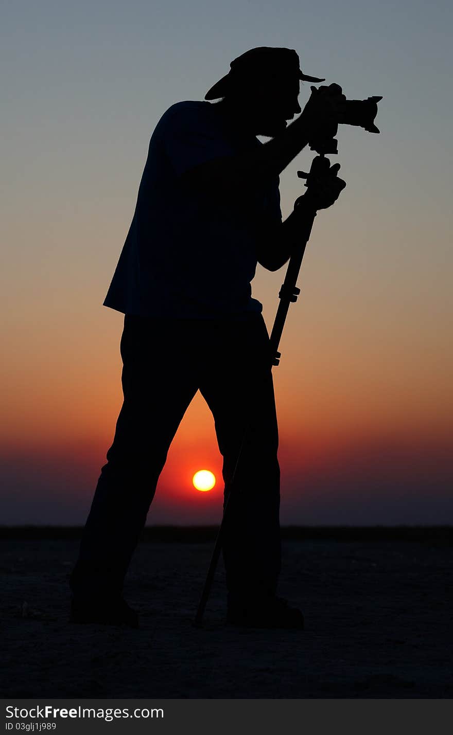 Silhouette of a young man in a hat taking pictures at sunset using a tripod. Silhouette of a young man in a hat taking pictures at sunset using a tripod