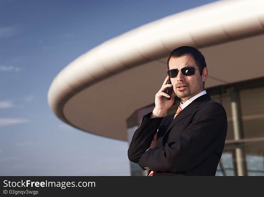 Close up of young cool business person in black suit and sunglasses being busy on cell phone with office building and blue sky in background. Close up of young cool business person in black suit and sunglasses being busy on cell phone with office building and blue sky in background.