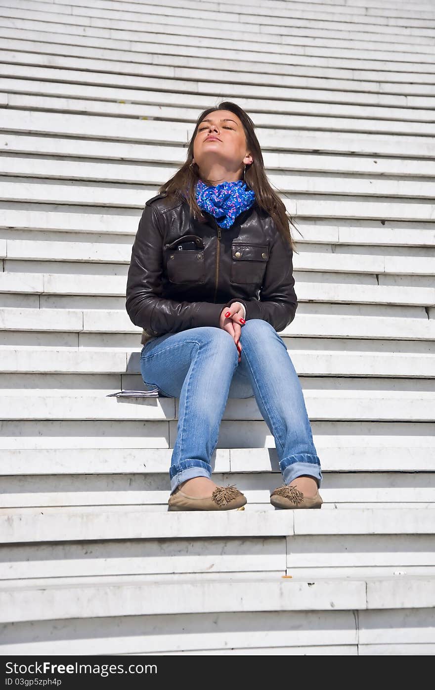 Attractive young woman sitting on the marble steps and tans. Sunlight. Eyes closed.