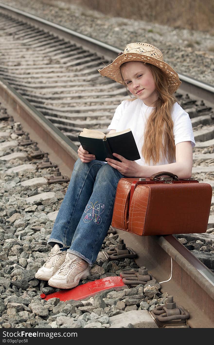 The young girl with a fair hair in a white scarf on a head with a brown sac sits on railway rails in expectation of a train and reads the book. The young girl with a fair hair in a white scarf on a head with a brown sac sits on railway rails in expectation of a train and reads the book