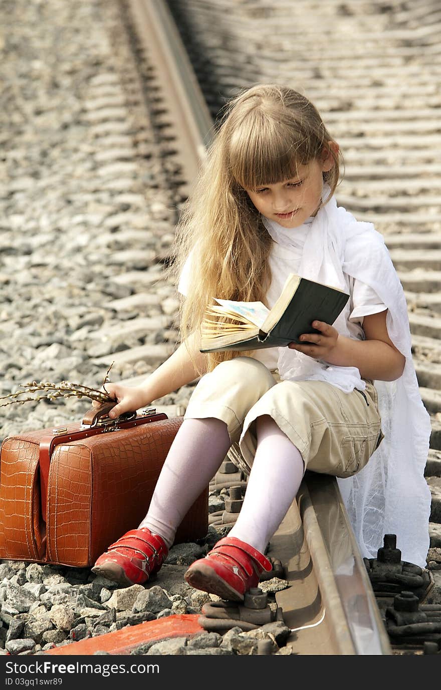 The young girl with a fair hair in a white scarf on a head  with a brown sac sits on railway rails in expectation of a train and reads the book. The young girl with a fair hair in a white scarf on a head  with a brown sac sits on railway rails in expectation of a train and reads the book