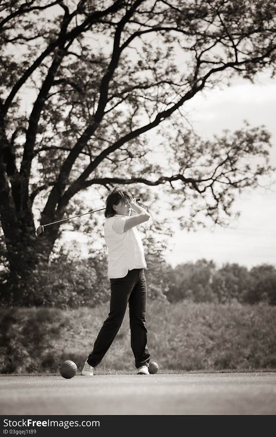 Active senior female golf player swinging golf club to tee off ball with beautiful tree in background, black & white conversion. Active senior female golf player swinging golf club to tee off ball with beautiful tree in background, black & white conversion.