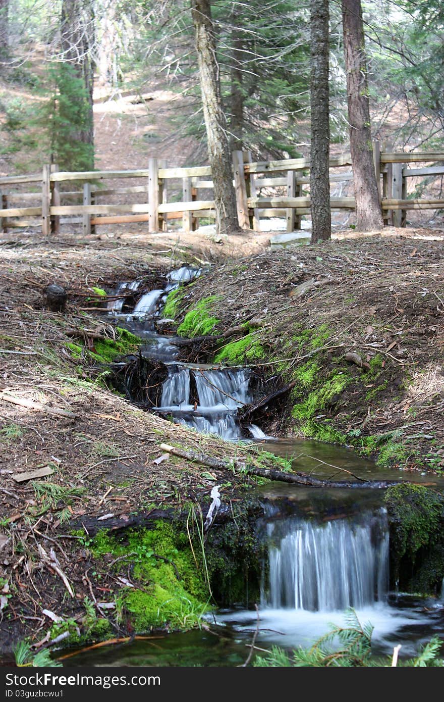 Small stream with waterfalls winding through hills and between trees under wooden fence. Small stream with waterfalls winding through hills and between trees under wooden fence