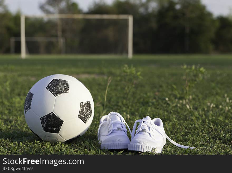 Old ball and white children's gym shoes on a football ground against gate. Old ball and white children's gym shoes on a football ground against gate