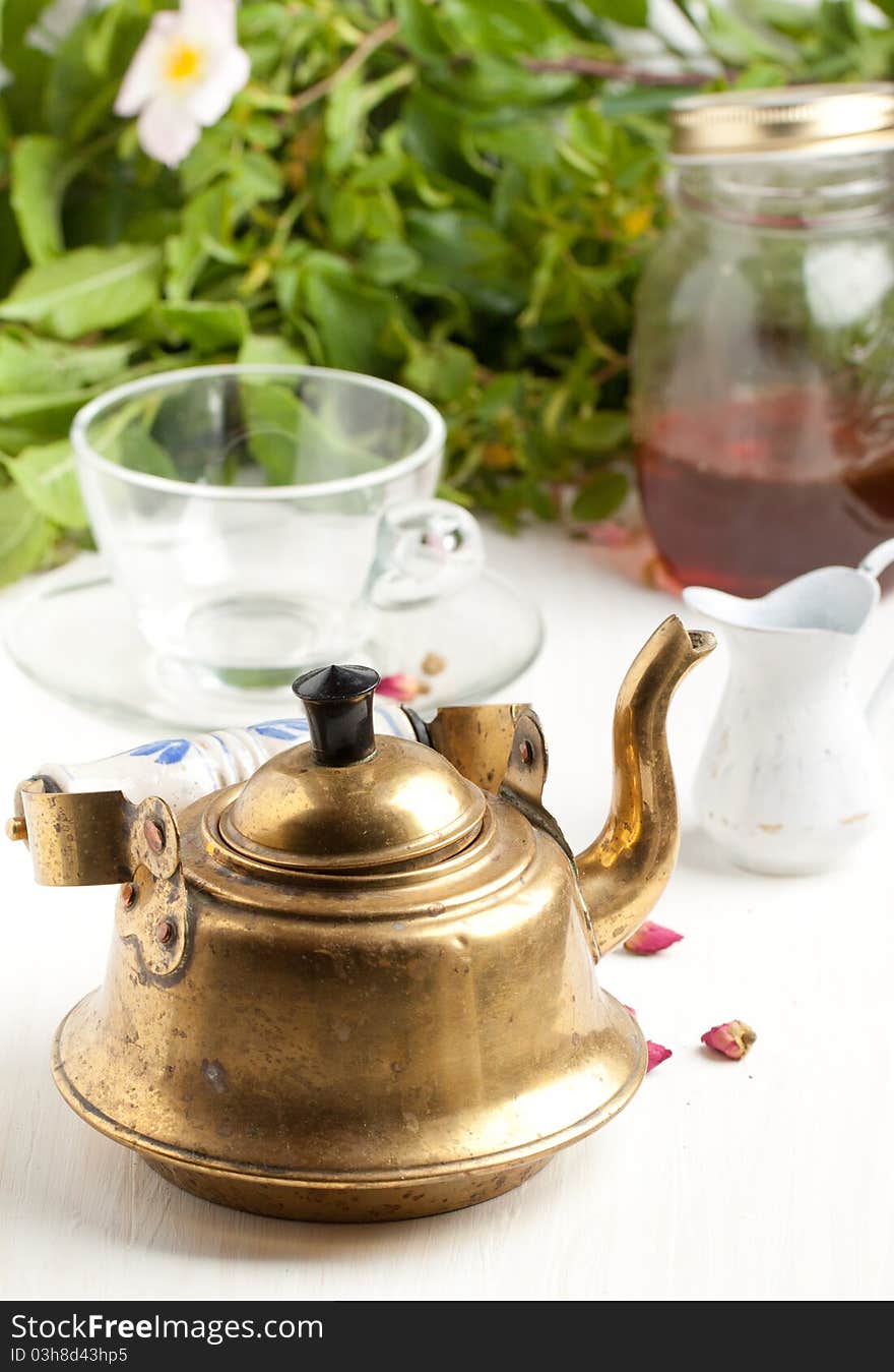 Old golden  teapot on white table with empty transparent teacup and little jug with green leafs as background. Old golden  teapot on white table with empty transparent teacup and little jug with green leafs as background