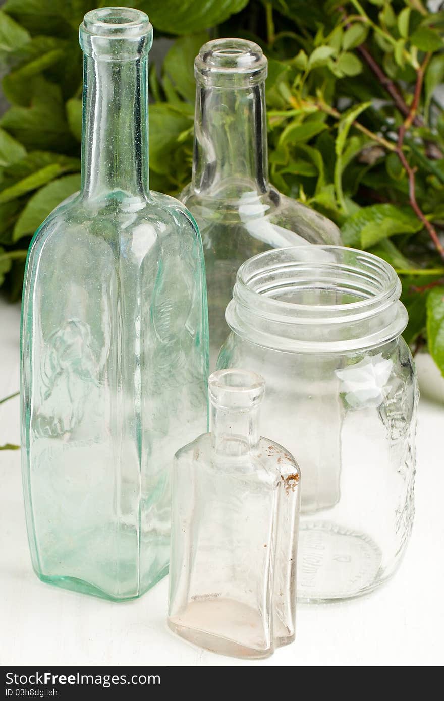 Composition with three vintage bottles and empty pot on white table with green leafs as background