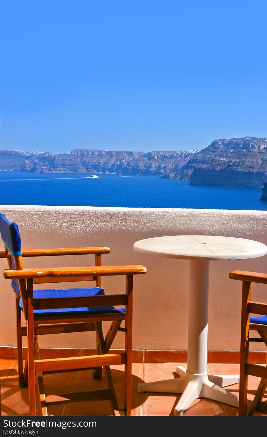 Blue chairs and a small table on beautiful terrace in Santorini with Caldera view and Aegean sea. As always clear blue sky in hot Greece. Great atmosphere for vacation and holidays. Blue chairs and a small table on beautiful terrace in Santorini with Caldera view and Aegean sea. As always clear blue sky in hot Greece. Great atmosphere for vacation and holidays.