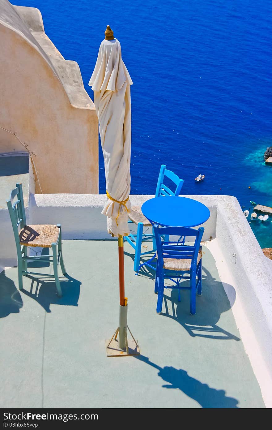 Blue chairs and a small table on beautiful terrace in Santorini with Caldera view and Aegean sea. As always clear blue sky in hot Greece. Great atmosphere for vacation and holidays. Blue chairs and a small table on beautiful terrace in Santorini with Caldera view and Aegean sea. As always clear blue sky in hot Greece. Great atmosphere for vacation and holidays.