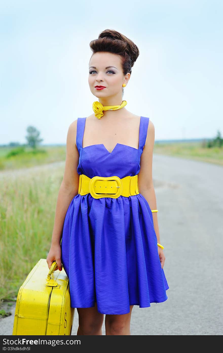 Beautiful smiling girl in a blue dress with a yellow suitcase on the road