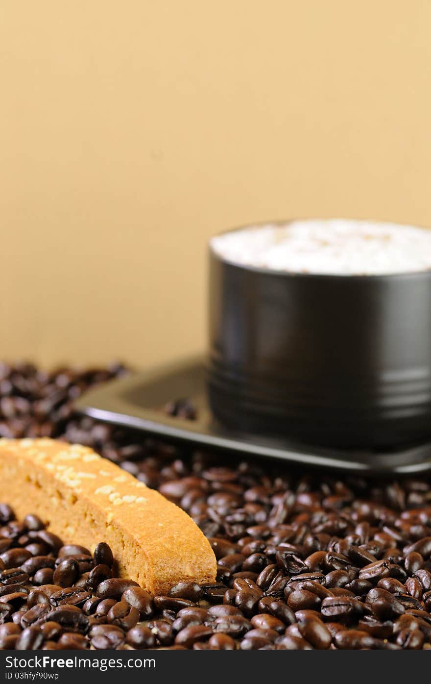 A creamy cappuccino in a black cup with a caramel biscotti on a bed of coffee beans with a shallow DOF. A creamy cappuccino in a black cup with a caramel biscotti on a bed of coffee beans with a shallow DOF.