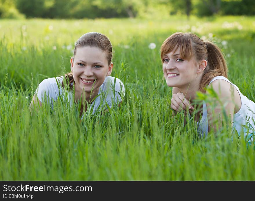 Two smiling girl lying in a grass and looking straight. They are wearing in a white t-shirt. Two smiling girl lying in a grass and looking straight. They are wearing in a white t-shirt