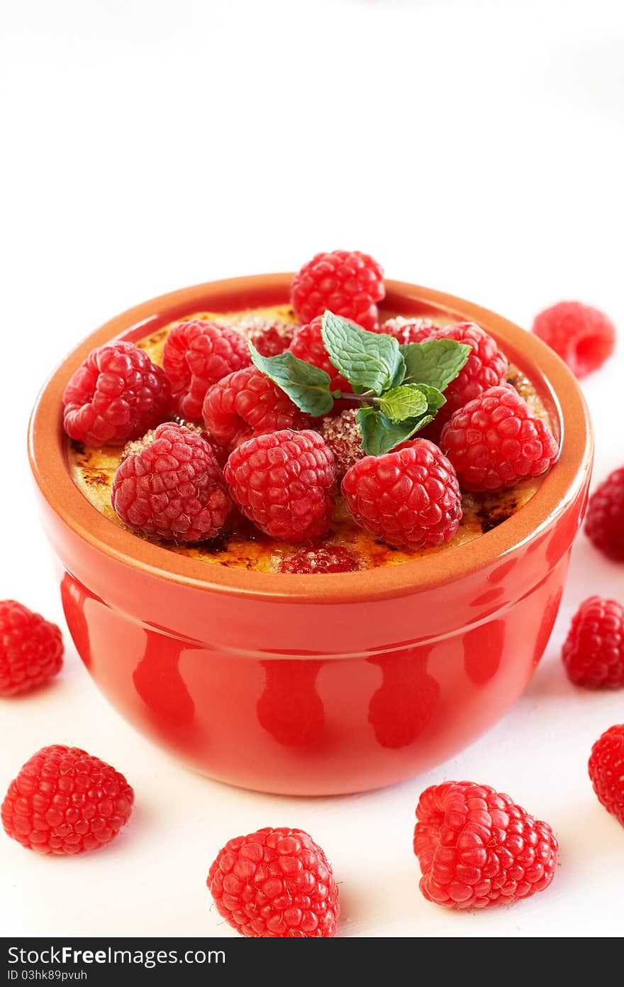 French creme brulee dessert with raspberries and mint covered with caramelized sugar in red terracotta ramekin on white background