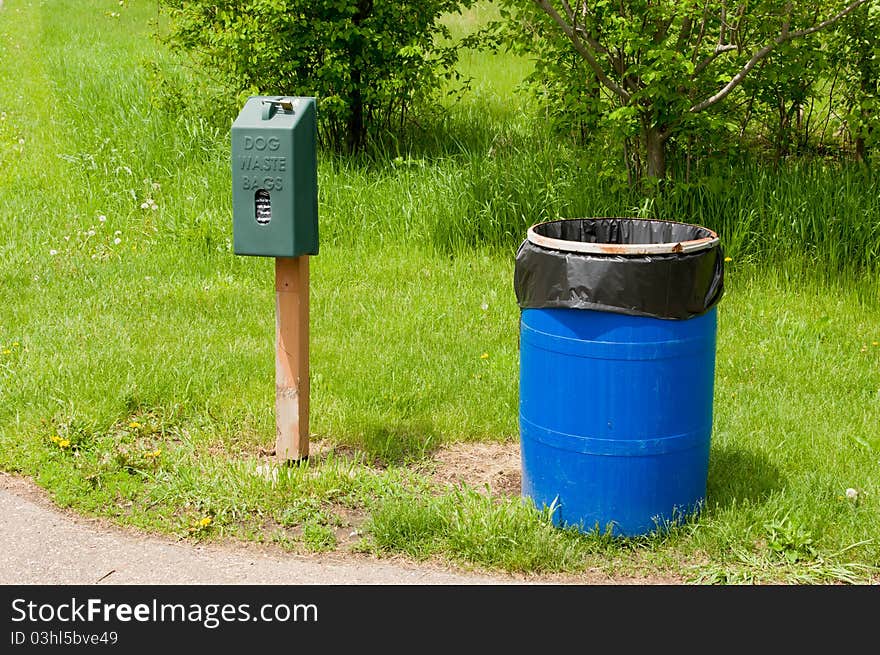 An Animal Waste Bag Dispenser by a Blue Trash Can in a Park. An Animal Waste Bag Dispenser by a Blue Trash Can in a Park