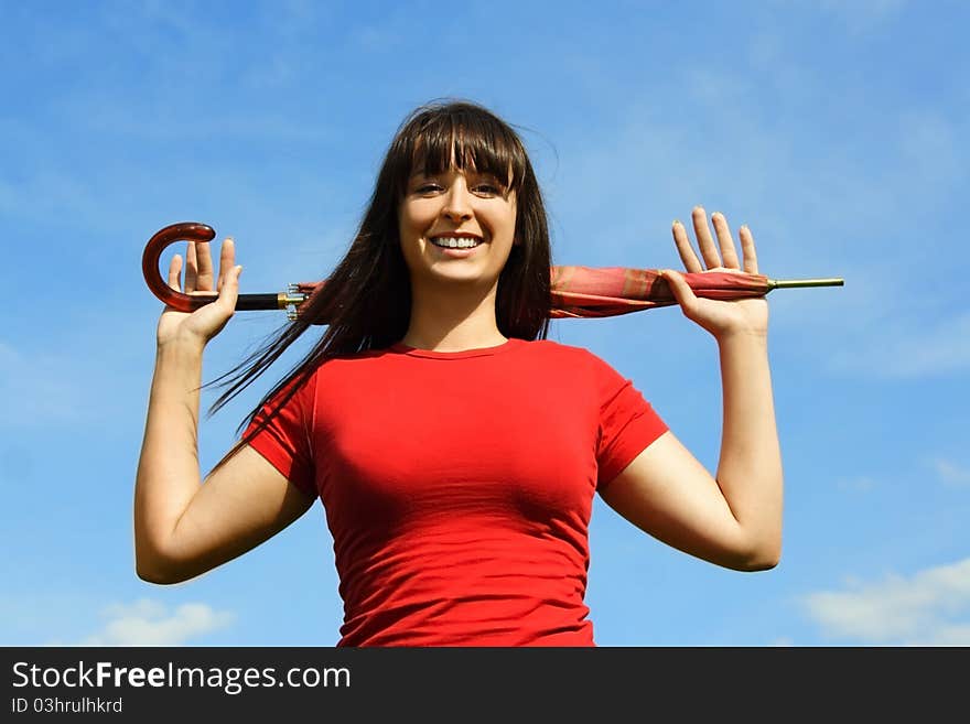 Young brunette girl in red shirt holding closed red umbrella behind head, blue sky. Young brunette girl in red shirt holding closed red umbrella behind head, blue sky