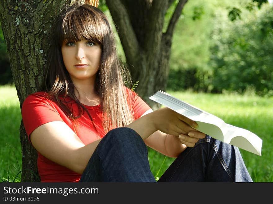 Young brunette girl in red shirt siting near tree and reading book, outdoor. Young brunette girl in red shirt siting near tree and reading book, outdoor