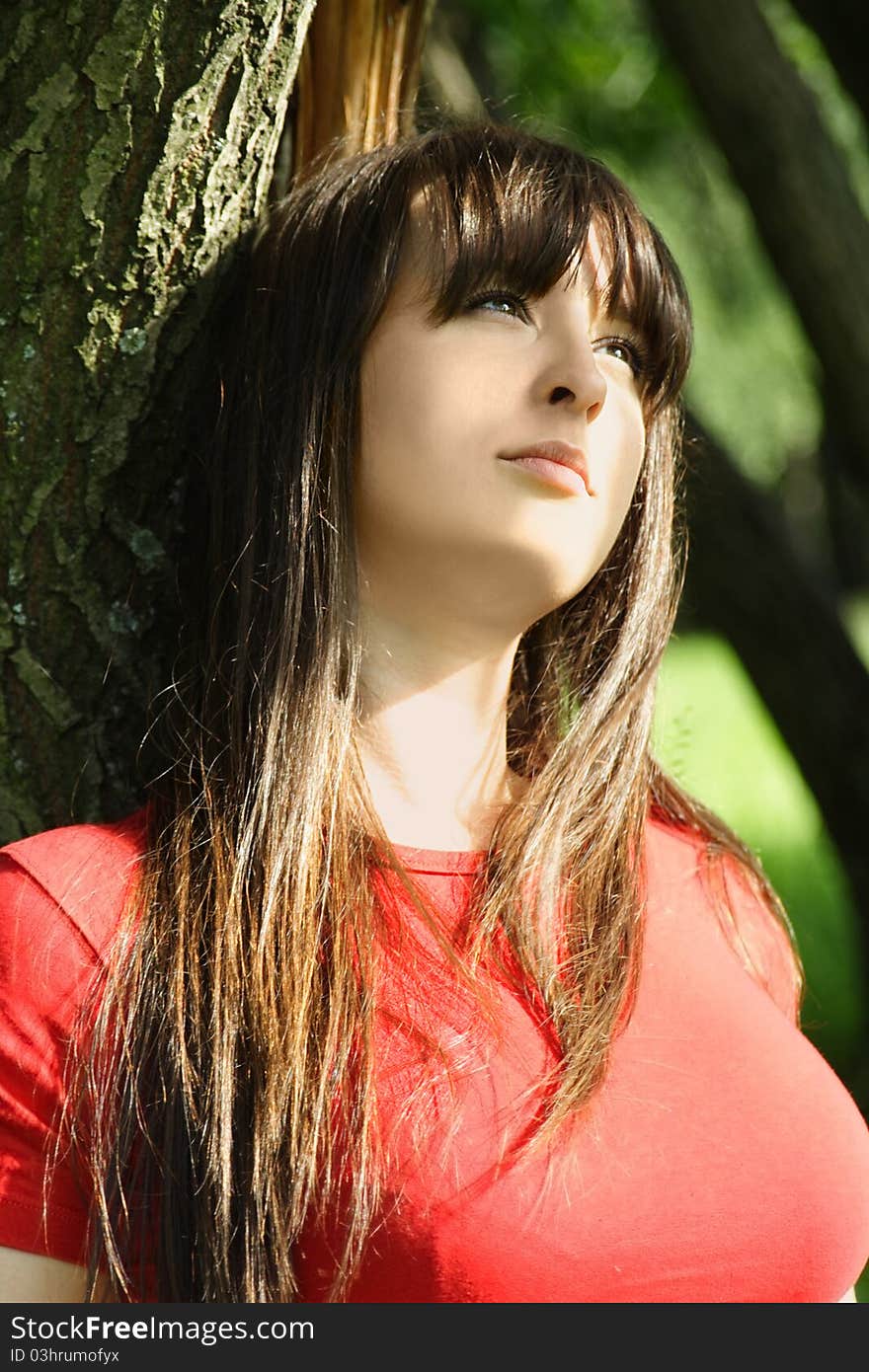 Young brunette girl in red shirt sitting near tree and looking at side. Young brunette girl in red shirt sitting near tree and looking at side