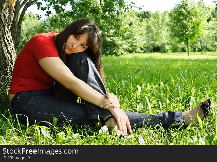 Young brunette girl in red shirt siting near tree and thinking, summer meadow