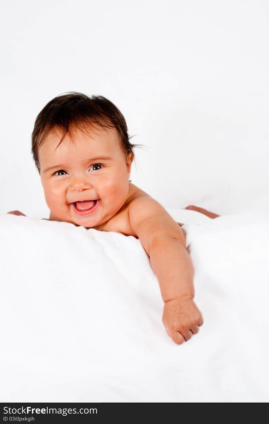 A mixed ethnicity baby smiles while laying on a white pillow and background.