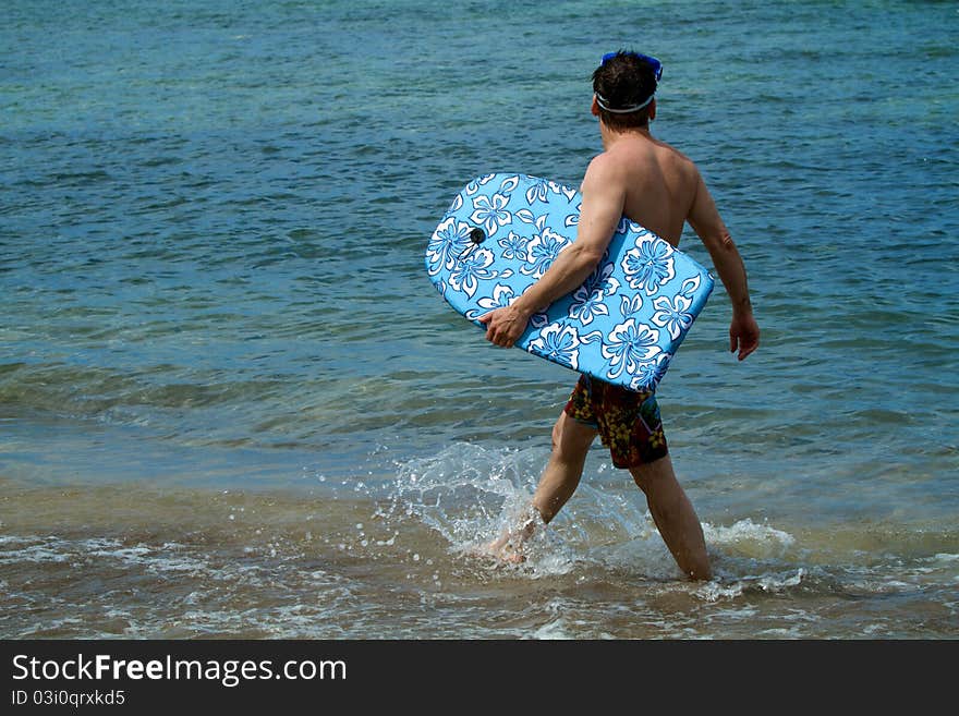 Man walks along the shore of a beach holding a bodyboard, looking for waves. He splashes water as he walks. Man walks along the shore of a beach holding a bodyboard, looking for waves. He splashes water as he walks.