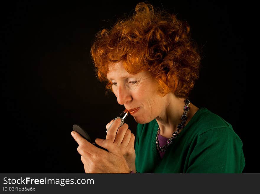 Curly red haired woman applies lipstick against a dark background. Curly red haired woman applies lipstick against a dark background