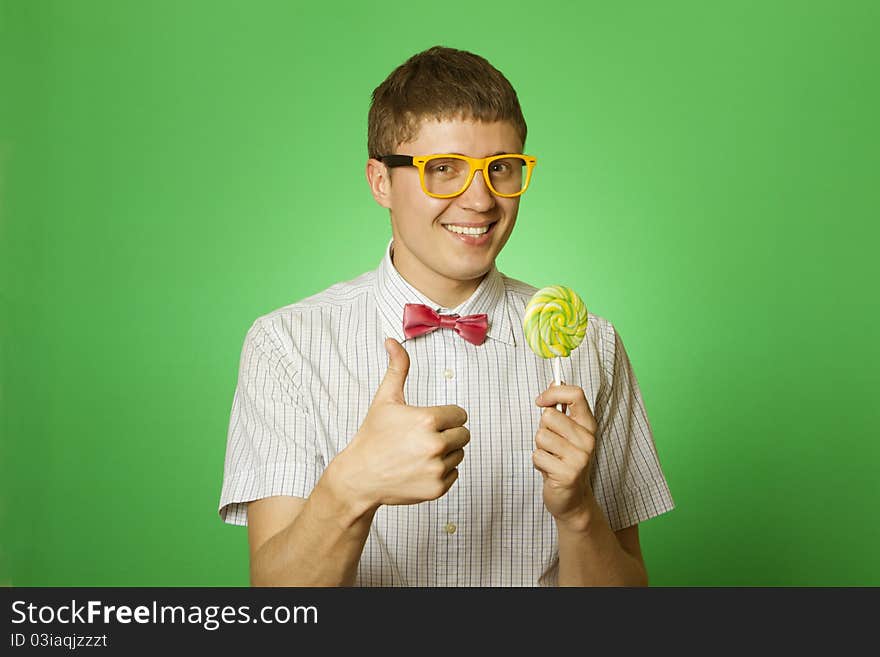 Closeup of an attractive young man in a shirt, bow tie and yellow sunglasses with a big lollipop. Thumbs up. Closeup of an attractive young man in a shirt, bow tie and yellow sunglasses with a big lollipop. Thumbs up