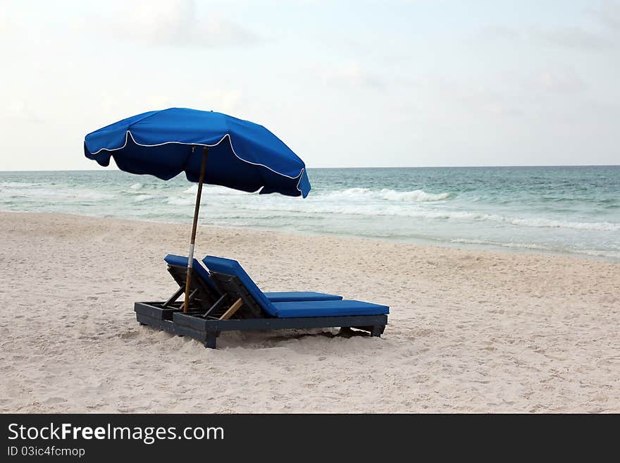 A lone pair of blue beach lounge chairs shaded by a matching umbrella with a view of the waves breaking on the shore. Horizontal shot. A lone pair of blue beach lounge chairs shaded by a matching umbrella with a view of the waves breaking on the shore. Horizontal shot.