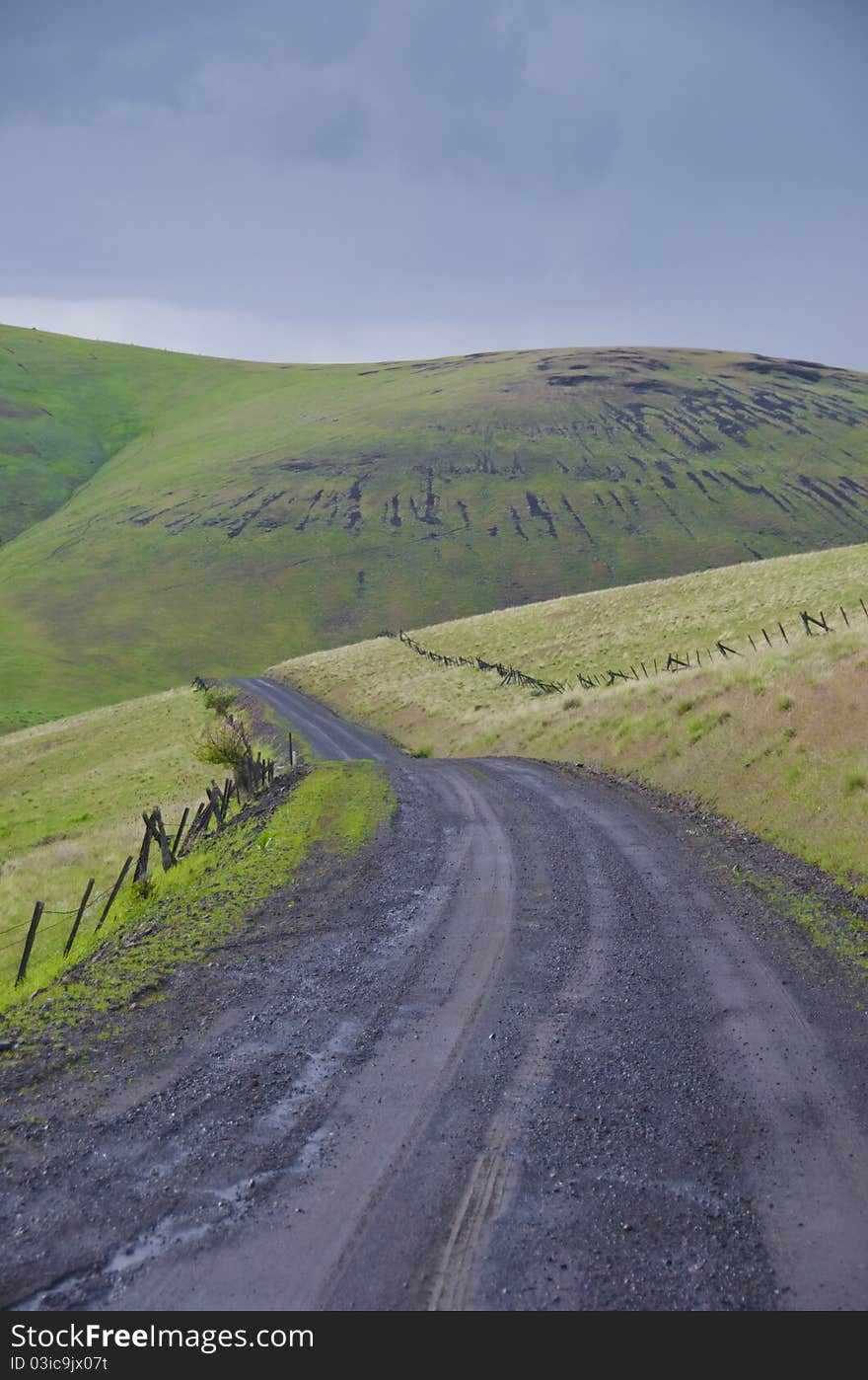 Black gravel road among grassy hills below a stormy sky. Black gravel road among grassy hills below a stormy sky