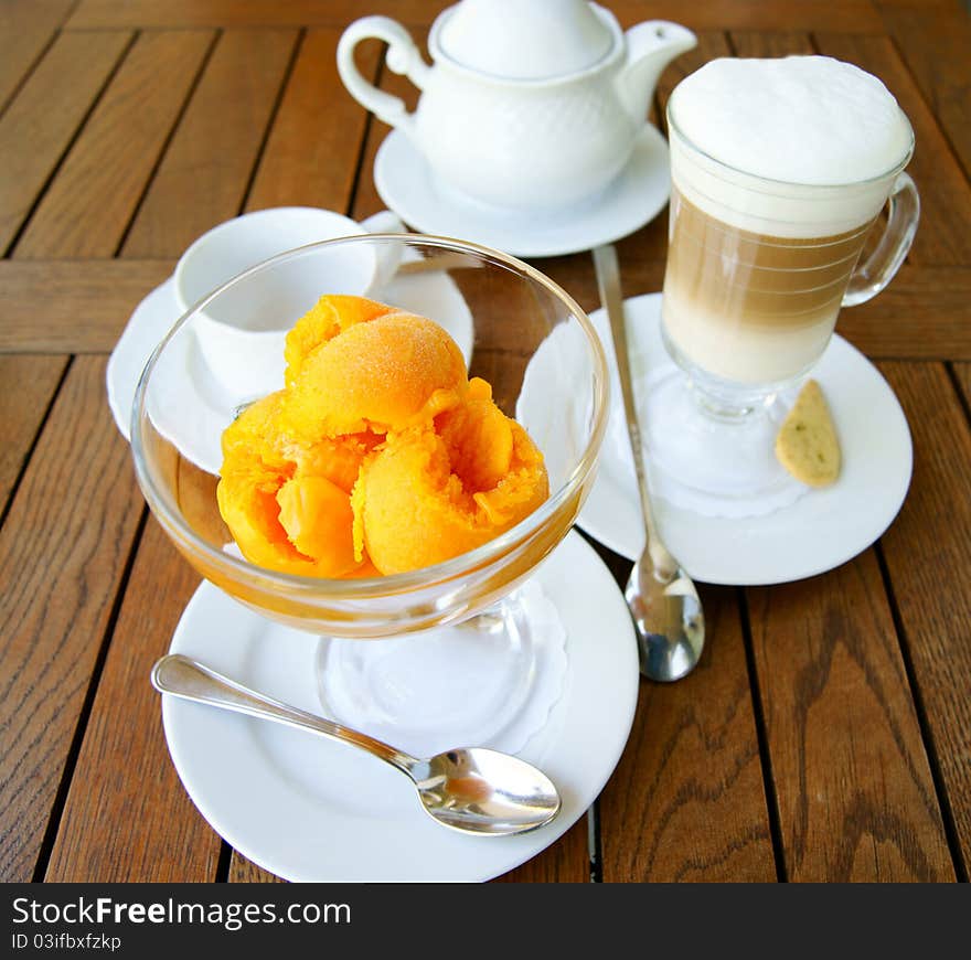 Orange ice cream in a glass bowl and glass of coffee latte  on the background of the wooden table surface. Orange ice cream in a glass bowl and glass of coffee latte  on the background of the wooden table surface