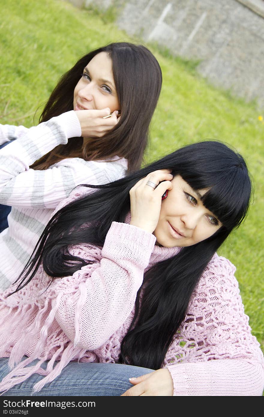 Attractive mother and teenage daughter talk on the phone outdoors in a park sitting back to back. Attractive mother and teenage daughter talk on the phone outdoors in a park sitting back to back