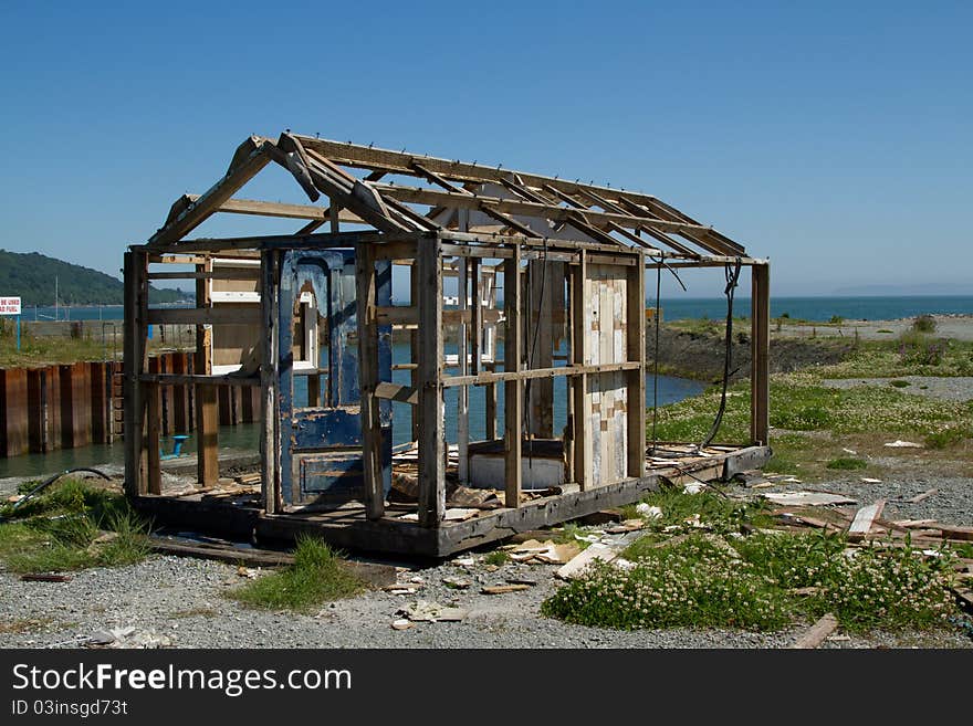 A wooden skeleton of an old hut, shed with blue door on waste ground with weeds against a blue sky. A wooden skeleton of an old hut, shed with blue door on waste ground with weeds against a blue sky.